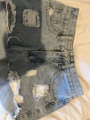 Ljusblåa slitna jeansshorts - Trendiga ljusblå jeansshorts med flera slitningar och råa kanter. Klassisk femficksmodell med knapp och dragkedja framtill. Perfekta för en avslappnad och cool stil.