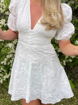 Vit kort klänning med puffärm från Adoore - Superfin vit kort klänning med puffärmar och v-ringning. Klänningen har broderade blommor, knappar framtill och markerad midja. Perfekt för sommarens alla tillfällen och ger en romantisk vibe.