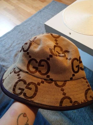 Beige bucket hat från Gucci - Säljer en beige bucket hat från Gucci med det klassiska GG-mönstret i brunt. Hatten har bred brätte och mörkbruna kanter. Materialet är canvasliknande och ger en cool streetwear-vibe. Perfekt för dig som vill sticka ut med en ikonisk accessoar.