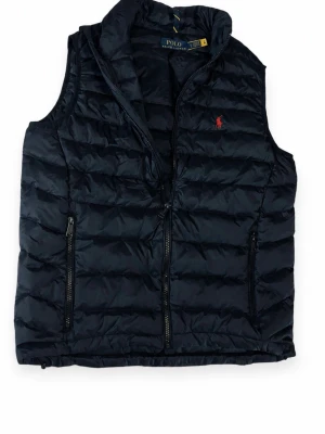 Svart dunväst från Polo Ralph Lauren - Snygg svart dunväst från Polo Ralph Lauren med quiltad design och dragkedja framtill. Västen har två praktiska fickor med dragkedja och en liten röd logga broderad på bröstet. Perfekt för lager-på-lager och streetstyle. Använd få gånger. Skriv för mer frågor 