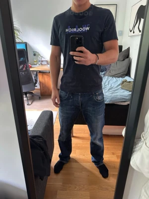 Svart t-shirt från Woolrich - Navy t-shirt från Woolrich med vit och blå logotyp på bröstet. Klassisk passform med rund hals och korta ärmar. Perfekt till jeans eller shorts för en avslappnad stil.