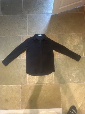 Mörkblå overshirt från Stone Island storlek 14y - Snygg mörkblå overshirt från Stone Island med klassisk krage, knappstängning och en bröstficka. Ikonisk logotyp på armen. Parfekt fom en chill sommaroutfitt