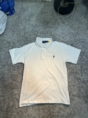 Vit pikétröja från Ralph Lauren - Klassisk vit pikétröja från Ralph Lauren med korta ärmar och krage. Tröjan har knappar framtill och den ikoniska broderade loggan i mörkblått på bröstet. Perfekt för en clean och stilren look.
