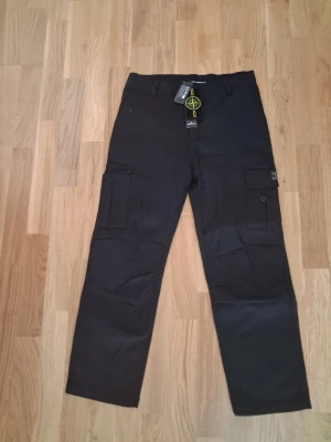 Svarta cargobyxor från Stone Island - Snygga svarta cargobyxor från Stone Island med klassiska cargofickor på sidorna och ikonisk logotyp på ena benet. Byxorna har raka ben, bälteshällor och är tillverkade i ett slitstarkt material som passar perfekt till en streetwear-look.
