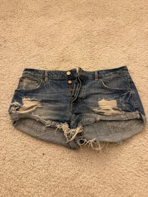 Blå jeansshorts med slitningar - Säljer ett par blå jeansshorts med råa kanter och slitna detaljer framtill. Shortsen har knappar i gylfen, klassiska fem fickor och en avslappnad passform. Perfekta för varma dagar och en cool streetstyle-look.