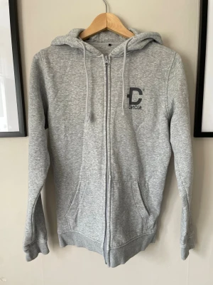 Grå zip-hoodie från DirtCult - Snygg gråmelerad hoodie från DirtCult med dragkedja, huva med dragsko och fickor fram. Tryck med DirtCult-logga på bröstet och stort tryck på ryggen med texten 'DirtCült San Diego'. Mjuk bomullsblandning och ribbade muddar vid ärmslut och nederkant.