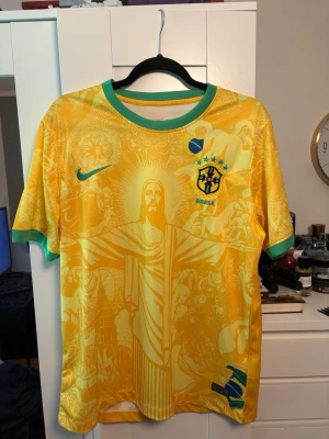 Brasilien fotbollströja Nike gul/grön - Aldrig använd gul Brasilien fotbollströja från Nike med gröna detaljer vid krage och ärmslut. Tryck med Kristusstatyn och brasilianska symboler på framsidan, samt Brasilien-emblem och Nike-logga. Tillverkad i lätt och ventilerande material, perfekt för fotbollsträning.