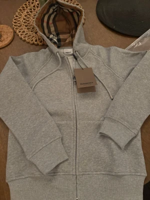 Grå zip hoodie från Burberry - Snygg grå zip hoodie från Burberry med klassiskt rutigt foder i huvan. Tillverkad i mjuk bomullsblandning, har dragkedja framtill och ribbade muddar vid ärmar och midja. Perfekt för en chill och stilren look. Passar S