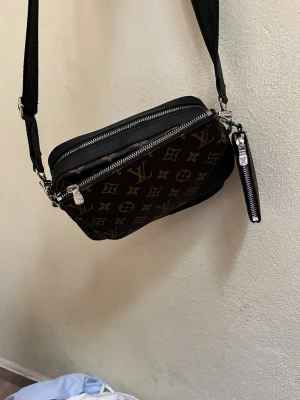 Louis Vuitton brun axelväska monogram - Snygg brun axelväska från Louis Vuitton med klassiskt monogrammönster i beige. Väskan har två fack med silverfärgade dragkedjor och en bred, justerbar axelrem. Tillverkad i tåligt konstläder och har en liten avtagbar ficka på sidan.