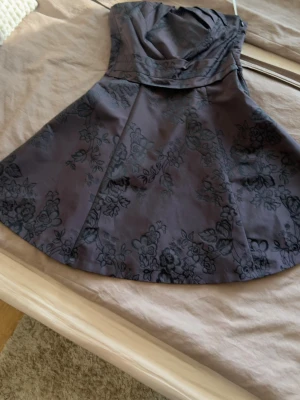Karen Millen festklänning - Snygg mörklila axelbandslös klänning med blommigt mönster och draperad överdel. Kjolen är utsvängd och klänningen har en elegant, kort längd. Perfekt för dig som vill sticka ut med en klassisk men trendig look.