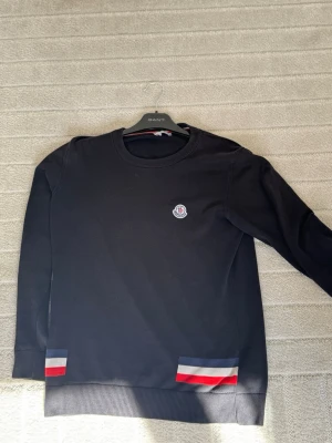Svart Moncler tröja med logga - Snygg svart tröja från Moncler med rund halsringning och diskret logga på bröstet. Tröjan har ribbade muddar och coola detaljer i rött, vitt och blått vid nederkanten. Perfekt för dig som gillar stilrena plagg med exklusiv känsla.