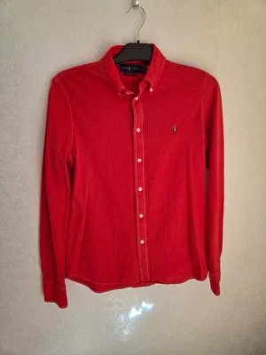 Röd skjorta från Ralph Lauren - Klassisk röd skjorta från Ralph Lauren med button-down krage och broderad logga på bröstet. Skjortan har långa ärmar och vita knappar framtill. Perfekt för en stilren och avslappnad look. Står storlek L, men den är mer som S. 