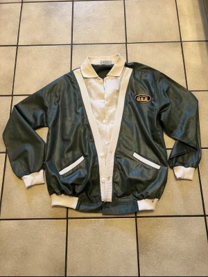 Grön bomberjacka med USA-brodyr storlek L/XL - Retro bomberjacka i mörkgrönt med vita detaljer och ribbade muddar. Framsidan har en broderad patch med texten 'California U.S.A.' på bröstet. Jackan har knappar framtill och två vita fickor. Materialet är glansigt och ger en vintagekänsla.