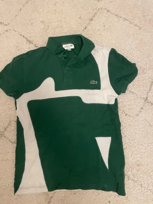Grön och vit pikétröja från Lacoste - Säljer en grön och vit pikétröja från Lacoste med coolt grafiskt mönster och klassisk krage. Tröjan har korta ärmar, knappar vid halsen och den ikoniska krokodilloggan på bröstet. Perfekt för dig som gillar sportig och stilren look.