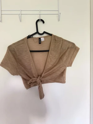 Beige croppad bolerotopp från H&M - Kort beige bolerotopp från Monki med knyt framtill. Toppen har korta ärmar och är gjord i ett tunt, lätt material som känns luftigt. Perfekt att styla över ett linne eller klänning för en trendig lager-på-lager-look.