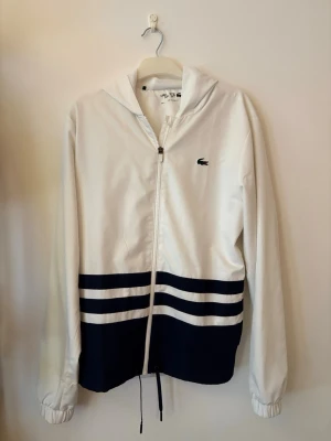 Lacoste vindjacka - Snygg vit vindjacka från Lacoste med marinblå detaljer och två horisontella ränder nedtill. Jackan har huva, dragkedja framtill, elastiska muddar och stor Lacoste-logga på ryggen. Perfekt för en sportig och stilren look.