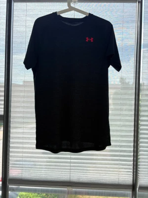 Svart t-shirt från Under Armour - Snygg svart t-shirt från Under Armour med diskret struktur i tyget och rött logotryck på bröstet. Klassisk rund halsringning och korta ärmar. Perfekt för dig som gillar sportig och enkel stil.