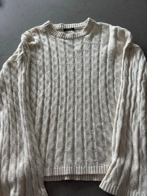 Vit kabelstickad tröja från Gina Tricot - Kabelstickad vit tröja från Gina Tricot med rund halsringning och ribbade muddar. Tröjan har ett klassiskt flätmönster och är perfekt för lager-på-lager-stil. Mjuk och skön känsla, passar till jeans eller kjol.