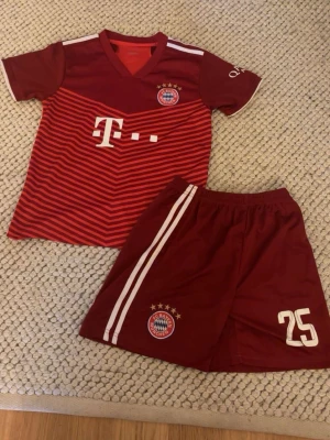 Bayern München matchställ Müller #25 - Snyggt rött matchställ från Bayern München med tröja och shorts. Tröjan har klubbmärke, sponsorlogga och Müller #25 på ryggen. Shortsen har vita ränder och klubbmärke. Tillverkat i lätt och ventilerande material, perfekt för fotbollsträning. Passar perfekt för pojkar som är 160-175