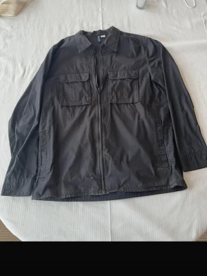 Svart overshirt från Divided - Stilren svart overshirt från Divided med dragkedja framtill och två stora bröstfickor med lock. Klassisk krage och raka ärmar ger en clean look. Tillverkad i bomullsmix som känns lätt och bekväm. Perfekt lager-på-lager-plagg för en avslappnad stil.