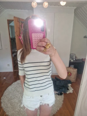 randig topp - Snygg vit topp med svarta horisontella ränder och rund hals. Modellen har korta ärmar och en mjuk, stickad känsla. Perfekt till jeans eller shorts för en fräsch look. Enkel och stilren design som passar till många outfits.