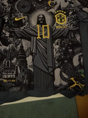 Nike Brasil fotbollströja svart/gul - Unik svart fotbollströja från Nike med Brasilien-tema och stort tryck av Kristusstatyn, gula detaljer och CBF-emblem. Tröjan har nummer 10 på bröstet och är tillverkad i Dri-FIT-material som andas. Perfekt för dig som vill sticka ut på planen.