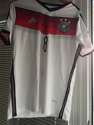 Tysklands matchtröja Özil #8 Adidas - Säljer en officiell Tyskland matchtröja från Adidas med Özil och nummer 8 på ryggen. Tröjan är vit med svarta och röda detaljer, har korta ärmar och tre svarta ränder på axlarna. Perfekt för fotbollsfans som vill ha en ikonisk landslagströja.