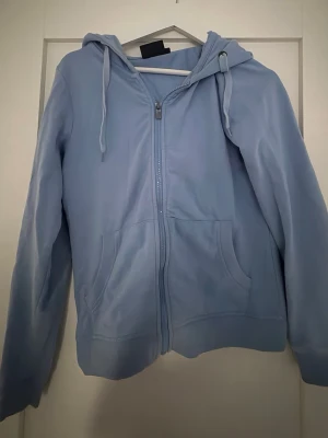 Ljusblå zip hoodie med huva - Ljusblå hoodie med dragkedja framtill och två stora fickor. Tröjan har en klassisk huva med dragsko och är tillverkad i mjukt material, perfekt för en avslappnad stil. Passar dig som gillar enkel och clean design.