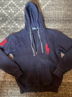 Marinblå hoodie från Polo Ralph Lauren - Snygg marinblå hoodie från Polo Ralph Lauren med dragkedja, gråa snören och stor röd logga på bröstet. På vänster ärm finns en röd siffra 3. Mjuk bomullskänsla och kängurufickor framtill. Perfekt för en avslappnad streetwear-look.bra skick. Storlek M