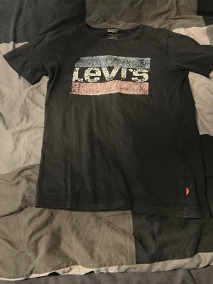 Svart Levi's t-shirt med logga - Säljer en svart t-shirt från Levi's med klassisk logga i blått, vitt och rött framtill. T-shirten har korta ärmar och rund halsringning. Tillverkad i mjuk bomull för skön känsla. Perfekt till jeans eller shorts.