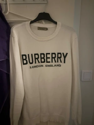 Vit Burberry tröja med logga - Snygg vit tröja från Burberry med stor svart logga och texten 'London England' på bröstet. Tröjan har rund halsringning, långa ärmar och ribbade muddar. Perfekt för dig som gillar klassisk stil med en modern twist.
