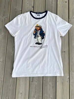 Vit Polo Bear t-shirt från Ralph Lauren - Vit t-shirt från Polo Ralph Lauren med den ikoniska Polo Bear-trycket på bröstet. T-shirten har mörkblå ribbad krage och är i slim fit-modell. Tillverkad i mjuk bomull och perfekt för en clean och klassisk stil.