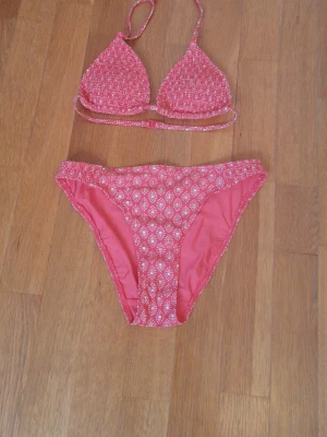 Rosa bikini med vitmönster - Söt rosa bikini med vita småmönster. Överdelen har trekantskupor och smala band som knyts i nacken och ryggen. Underdelen har samma mönster och klassisk passform. Perfekt för strandhäng och pooldagar.