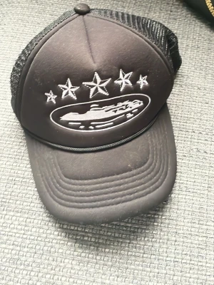Svart truckerkeps med broderi - Säljer en svart truckerkeps med mesh på sidorna och bak. Framtill finns ett broderat motiv med fem stjärnor och ett ovalt märke med ett fordon. Kepsen har böjd skärm och justerbar snapback. Perfekt för dig som gillar streetwear.