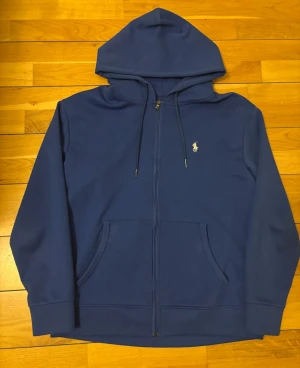 Blå hoodie från Polo Ralph Lauren - Snygg blå hoodie från Polo Ralph Lauren med dragkedja, huva med dragsko och kängurufickor. Klassisk vit logga broderad på bröstet. Tillverkad i mjukt bomullsmaterial som är perfekt för chill dagar eller när du vill ha en clean look.