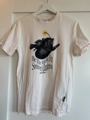 Vit t-shirt med tryck från Dedicated - Vit t-shirt från Dedicated i ekologisk bomull med tryck av en djuphavsfisk och texten 'Be The Light In Someone's Darkness'. Klassisk passform och rund hals. Har för mig att nypris var 400 kr, säljer för 200 kr