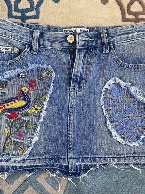 Broderad jeanskjol från Feurde Jeans - Cool kort jeanskjol med broderade detaljer från Feurde Jeans. Framsidan har färgglada broderier med fågel och blommor samt blått mönster, medan baksidan har en patch med flygplansmotiv. Rå kant nertill för en avslappnad look.