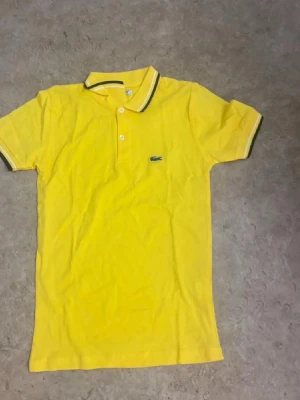 Gul pikétröja från Lacoste - Snygg gul pikétröja från Lacoste med klassisk krage och två knappar framtill. Tröjan har korta ärmar med kontrastfärgade detaljer på krage och ärmslut samt den ikoniska krokodilloggan på bröstet. Perfekt för en fräsch och sportig look.