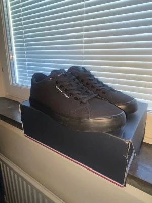 Svarta sneakers Tommy Hilfiger - Snygga svarta sneakers från Tommy Hilfiger med lågt skaft och klassisk design. Skorna har ovandel i textil, svarta skosnören och diskret logga på sidan. Yttersulan är svart med gummisula undertill för bra grepp. Perfekta till jeans eller shorts.