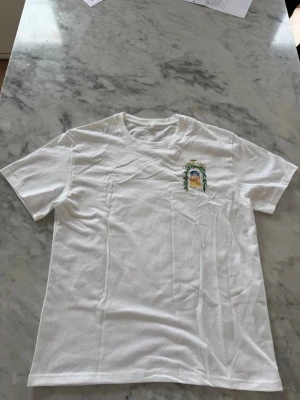 Vit t-shirt Casablanca tryck - Vit t-shirt från Casablanca med ett färgglatt tryck på bröstet som föreställer en byggnad och texten 'Avenida Casablanca'. T-shirten har rund hals och korta ärmar. Perfekt för dig som gillar stilrena plagg med unika detaljer.