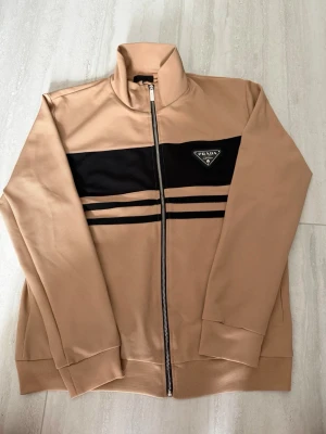 Beige zip hoodie från Prada - Snygg beige hoodie från Prada med svart panel och två svarta ränder över bröstet. Dragkedja framtill och hög krage. Ikonisk Prada-logga i svart och guld på bröstet. Tillverkad i mjukt material som känns lyxigt och bekvämt.