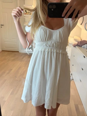 Vit kort sommarklänning med puffärm - Söt vit kort klänning med puffärmar och markerad midja. Klänningen har en lätt rynkad byst, rundad urringning och knytband vid ärmarna. Perfekt för varma dagar och har en luftig, flowy passform.
