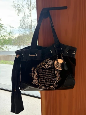 Juicy Couture svart sammetshandväska och nästan helt oanvänd  - Juicy Couture handväska i svart sammet med detaljer i svart läder. Väskan har tryckt text och dekorativa emblem på framsidan, samt en stor metallberlock och en tygrosett på sidan. Två handtag och hjärtformat J-märke på baksidan. Perfekt för dig som gillar statementväskor.