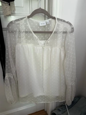 Vit prickig blus från Vila - Söt vit blus från Vila med transparenta långärmar och små prickar i tyget. Blusen har spetsdetaljer vid axlarna och knytband vid halsen. Perfekt för en trendig och romantisk look.