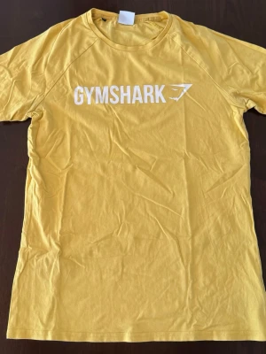 Gymshark t-shirt  - Snygg gul t-shirt från Gymshark med vit logga tryckt på bröstet. Klassisk rund halsringning och korta ärmar. Tillverkad i mjuk bomull som känns skön mot huden. Perfekt för dig som gillar sportiga och stilrena plagg.