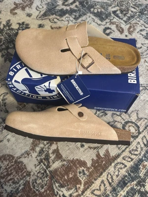  Birkenstock boston bs ’sand’ - Stilrena beige tofflor från Birkenstock i mjuk mocka med justerbart spänne i metall. Klassisk slip-in modell med präglad logga på sidan och bekväm fotbädd i kork. Perfekta för en avslappnad och trendig look hemma eller ute.
