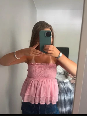 Rosa bandeau topp med volanger - Säljer en söt rosa bandeau topp med smockad överdel och dubbla lager volanger nedtill. Toppen är axelbandslös och har en luftig, flowy passform. Perfekt till sommaren och enkel att styla med jeans eller kjol.