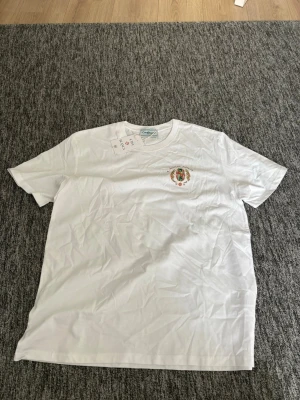 Vit Casablanca Tennis Club t-shirt - Vit t-shirt från Casablanca med färgglatt tryck på bröstet och stort broderat motiv på ryggen med texten 'Casablanca Tennis Club'. Klassisk passform och rund hals. Perfekt för dig som gillar stilrena men ändå unika plagg.