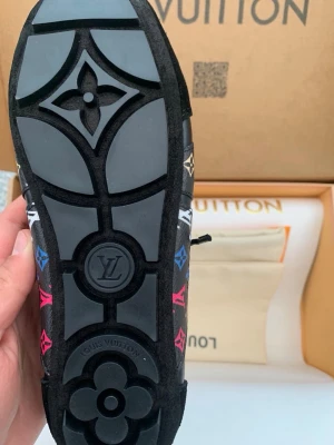 Louis Vuitton svarta sneakers med monogram - Svarta sneakers från Louis Vuitton med färgglada monogramdetaljer i blått, rött, grönt och vitt. Skon har en svart sula med LV-logga och blommönster, samt svarta skosnören och detaljer i läder och mocka. Perfekt för dig som gillar lyxiga och unika sneakers.