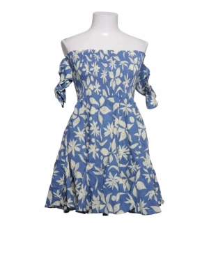 Blå offshoulder blommig klänning - Supersöt blå kortklänning med vita blommor och offshoulder-design. Klänningen har smock upptill, knytband på ärmarna och en luftig kjol. Perfekt för varma dagar och ger en riktigt somrig vibe!